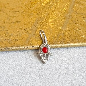 Könnte beinhalten: Silberner Anhänger in Form einer Hamsa-Hand mit roter Mitte. Die Hamsa ist ein beliebtes Symbol für Schutz und Glück.