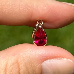 May include: A small, teardrop-shaped pendant with a vibrant red gemstone set in a gold-colored metal. The pendant is held between two fingers against a blurred green background, showcasing its size and color.