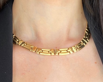 Könnte beinhalten: Eine goldene Kettenkette mit einem klobigen, geometrischen Gliederdesign. Die Kette wird von einer Person mit langen dunklen Haaren getragen.