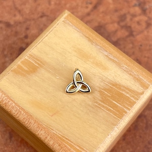 Può includere: Un ciondolo a nodo celtico dorato a forma di triangolo. Il ciondolo è su una superficie di legno.