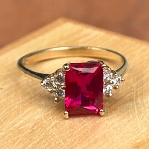 May include: A gold ring with a large, rectangular, red ruby gemstone surrounded by smaller, clear gemstones.
