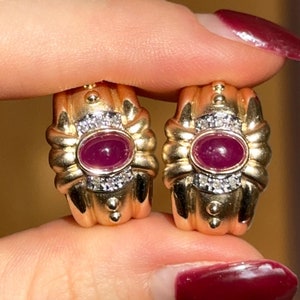May include: A pair of gold hoop earrings with a ruby and diamond accent. The earrings are held in a hand with red nail polish.