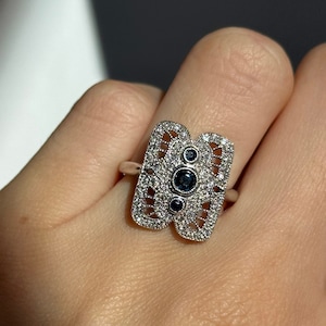 May include: A silver ring with a geometric design featuring three blue gemstones surrounded by smaller white gemstones.