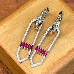 May include: A pair of silver-toned earrings with a rectangular design. The earrings are encrusted with small, clear gemstones and feature a horizontal band of red gemstones. The earrings are displayed on a wooden box.