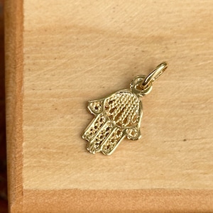 Könnte beinhalten: Ein goldfarbener Hamsa-Hand-Anhänger. Der Anhänger ist mit einem filigranen Design verziert.