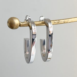 May include: A pair of silver hoop earrings with a slightly squared shape. The earrings are hanging from a gold stand.