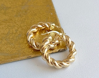 May include: A pair of gold hoop earrings with a twisted rope design. The earrings are on a white surface.