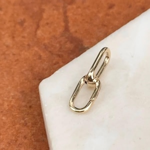 May include: A small, gold-colored jewelry link with an oval and a rectangular shape. The link is shown on a white marble surface with a brown background.