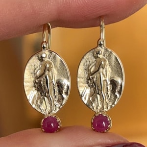 Op de afbeelding: Gouden oorbellen met een gedetailleerd figuurontwerp en een roze edelsteenhanger.