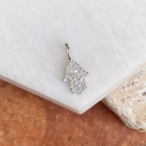 Könnte beinhalten: Ein kleiner, silberfarbener Hamsa-Hand-Anhänger, besetzt mit zahlreichen kleinen, klaren Edelsteinen. Der Anhänger hängt an einer kleinen Schlaufe. Der Hintergrund ist eine weiße Marmoroberfläche mit einem beigefarbenen Abschnitt.