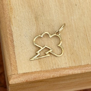 May include: Gold charm in the shape of a cloud with a lightning bolt inside. The charm is on a wooden surface.