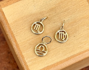 May include: Three gold-colored zodiac sign charms, each featuring a different astrological symbol: Libra, Virgo, and Scorpio. The charms are displayed on a wooden surface.