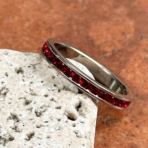 May include: A silver-toned metal eternity band ring featuring a continuous channel setting of small, vibrant red gemstones that wrap around the entire circumference of the band, creating a uniform and polished appearance with a smooth metallic finish.