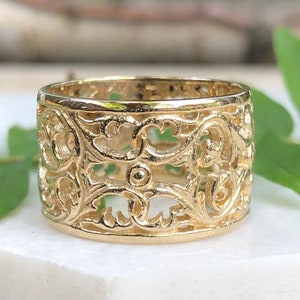 Può includere: Un anello in filigrana d'oro con un design delicato e intricato. L'anello è largo e ha una fascia arrotondata.