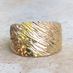 May include: A gold band ring with a textured, diagonal pattern. The ring is wide and has a rounded shape.