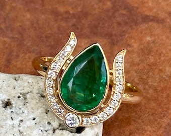 Könnte beinhalten: Ein Goldring mit einem zentralen birnenförmigen grünen Smaragd, der von einem geschwungenen Heiligenschein aus kleinen weißen Diamanten umgeben ist. Die Fassung besteht aus Gelbgold, wobei die Diamanten eine Halbmondform um den Smaragd bilden.