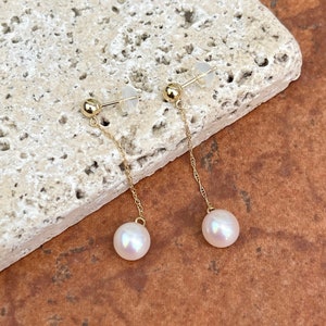 May include: A pair of gold drop earrings with white pearls. The earrings have a small gold ball at the top and a delicate gold chain that hangs down to a pearl.