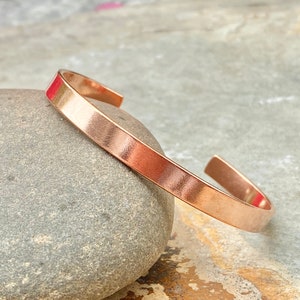 May include: A simple copper bracelet with a smooth, polished finish. The bracelet is open ended and has a slightly curved shape.