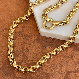 May include: A gold-colored chain necklace with a rounded link design. The necklace has a lobster clasp closure. The jewelry is displayed on a white surface against a brown background.