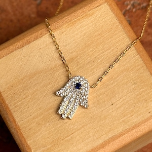 Könnte beinhalten: Eine goldene Kettenkette mit einem Hamsa-Hand-Anhänger. Der Anhänger ist mit klaren Zirkonia-Steinen besetzt und hat ein blaues böses Auge in der Mitte.