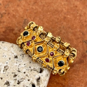 Könnte beinhalten: Ein goldener Ring mit einem geflochtenen Design und schwarzen und roten Edelsteinen. Der Ring liegt auf einem hellbraunen Stein.