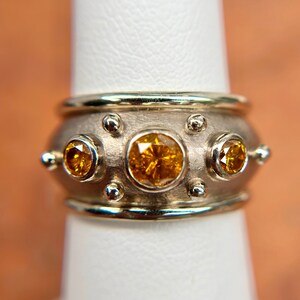 May include: A silver ring with three large, round, yellow-orange gemstones. The ring has a wide band with smaller silver accents and a gold-colored trim. The gemstones are set in a bezel setting, and the ring is displayed on a white surface.