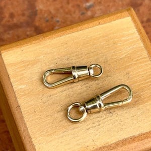 May include: Two gold-toned metal lobster claw clasps, each featuring a spring-loaded gate mechanism and a small circular swivel loop at the base, designed for attaching to jewelry chains or other hardware components for secure fastening.