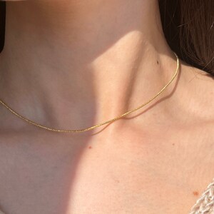 May include: A delicate gold necklace with a textured, beaded appearance. The necklace is shown against a person's skin, highlighting its simple elegance. The chain is thin and appears to be a choker style.