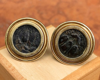 May include: A pair of gold-toned cufflinks featuring dark, circular insets with detailed, antique designs. The cufflinks are set on a wooden surface, showcasing their intricate craftsmanship and historical aesthetic.