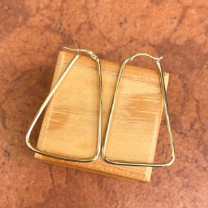 May include: A pair of gold triangle-shaped hoop earrings on a wooden surface.