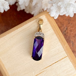 Könnte beinhalten: Ein rechteckiger Anhänger mit einem facettierten, tiefvioletten Edelstein. Der Anhänger hat eine goldfarbene Öse und Akzente, mit kleinen, klaren Steinen oben. Der Edelstein ist etwa 2,5 cm lang.