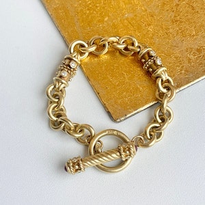 Könnte beinhalten: Ein goldenes Kettenarmband mit einem Schiebeverschluss. Die Kette ist mit kleinen Diamanten und einem roten Stein verziert.