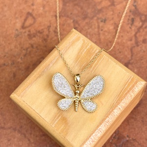 May include: A gold-colored butterfly pendant necklace. The butterfly has white, sparkling wings and a detailed body. The necklace is displayed on a wooden box against a brown background. The chain is thin and gold.