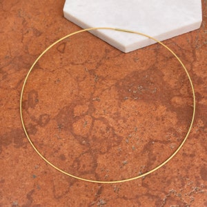 Op de afbeelding: Een goudkleurige ketting met een eenvoudig, cirkelvormig ontwerp. De ketting is gemaakt van een dun, flexibel materiaal en heeft een sluiting aan de achterkant. De ketting wordt gepresenteerd op een bruin en wit oppervlak.