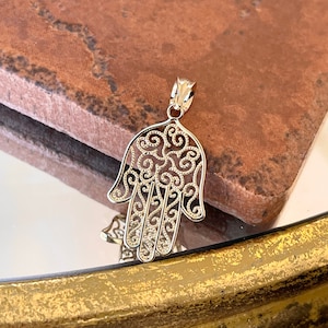Könnte beinhalten: Goldfarbener Hamsa-Hand-Anhänger mit aufwendigem filigranem Design. Der Anhänger weist ein detailliertes Muster aus Wirbeln und Kurven innerhalb der Handform auf. Der Anhänger wird auf einer reflektierenden Oberfläche mit verschwommenem Hintergrund präsentiert.