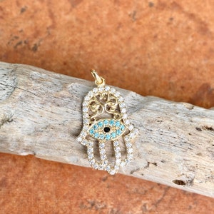 Könnte beinhalten: Goldfarbener Hamsa-Hand-Anhänger mit einem blauen bösen Augen-Anhänger und klaren Kristall-Akzenten.