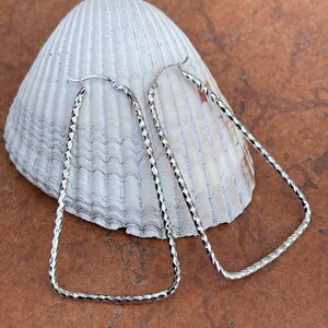 May include: A pair of silver, geometric, open hoop earrings. The earrings are made of a textured metal and have a rectangular shape.