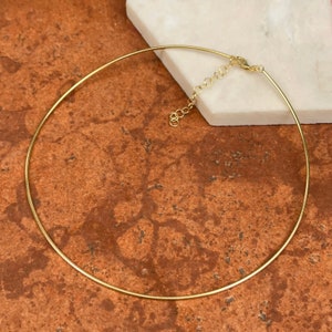 May include: A gold-colored choker necklace with an adjustable clasp. The necklace is a thin, smooth, and circular design. The clasp and adjustment chain are also gold-toned. The necklace is displayed on a white surface against a brown background.