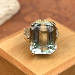 May include: A gold ring featuring a large, rectangular, light blue gemstone. The ring has intricate detailing on the band and around the gemstone setting. The gemstone is clear with a slight tint of blue and brown.