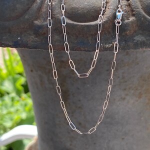 Pu&ograve; includere: Una collana a catena d'argento con un delicato design a maglie rettangolari. La catena &egrave; mostrata drappeggiata su una superficie testurizzata.