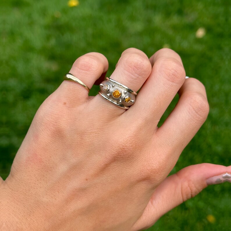 May include: A silver ring with three round, yellow-orange gemstones and a textured band. A smaller gold ring is also visible. The rings are displayed on a hand against a blurred green background.
