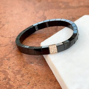 May include: A black bracelet with a rose gold square charm set with clear cubic zirconia stones.