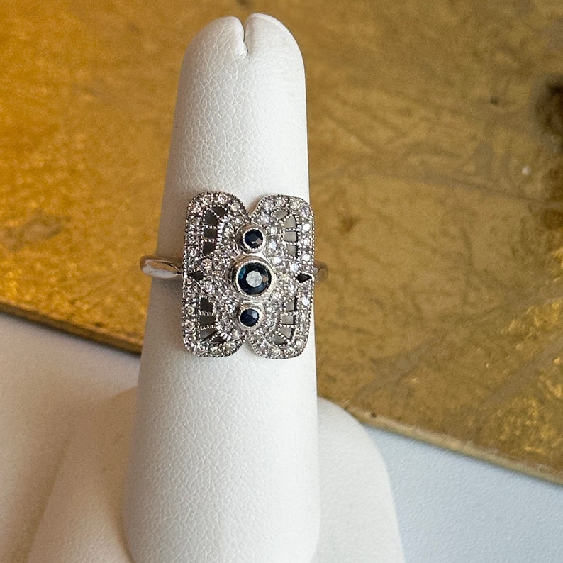 May include: A silver ring with a filigree design featuring three blue sapphires and smaller white diamonds.