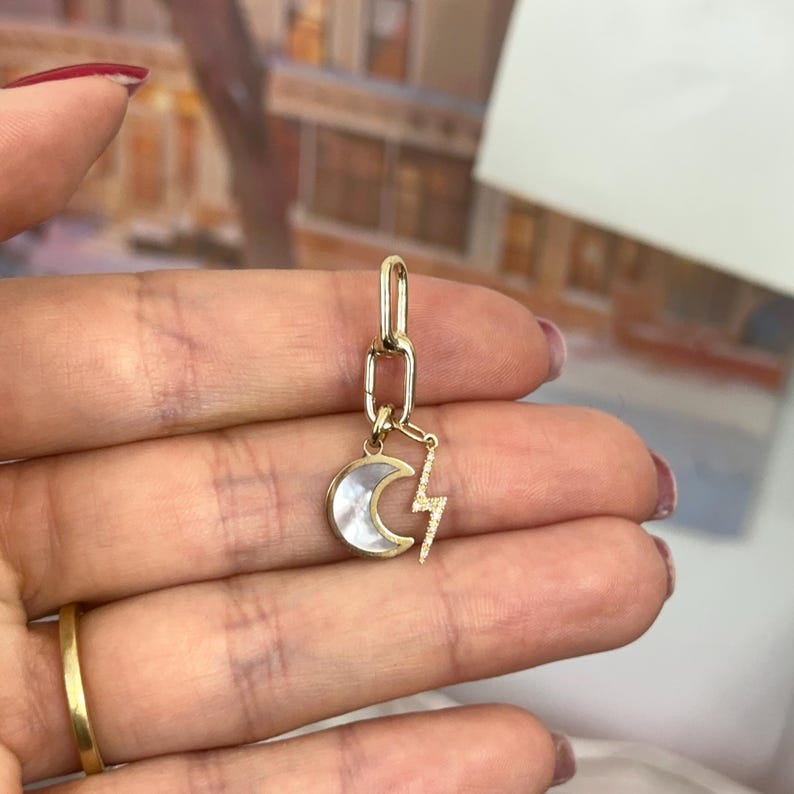 May include: A gold-colored charm featuring a crescent moon with a white inlay and a lightning bolt pendant. The charm is attached to a gold-colored chain link. The jewelry is held in a hand with a gold ring.