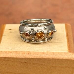 May include: A silver ring with a wide band, featuring three large, round, amber-colored gemstones. Smaller silver spheres and gemstones are set around the band. The ring is displayed on a wooden surface.