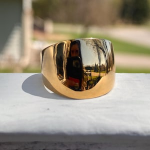 May include: A gold-colored ring with a wide band and a smooth, polished surface. The ring reflects the surrounding environment, including trees and a person holding a phone. The ring is a statement piece.