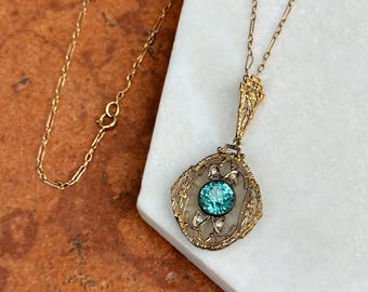 Op de afbeelding: Een goudkleurige hangerketting met een sierlijk filigraanontwerp. De hanger heeft een centrale, ronde, turquoisekleurige steen omringd door kleinere, heldere stenen. De ketting is delicaat en goudkleurig.