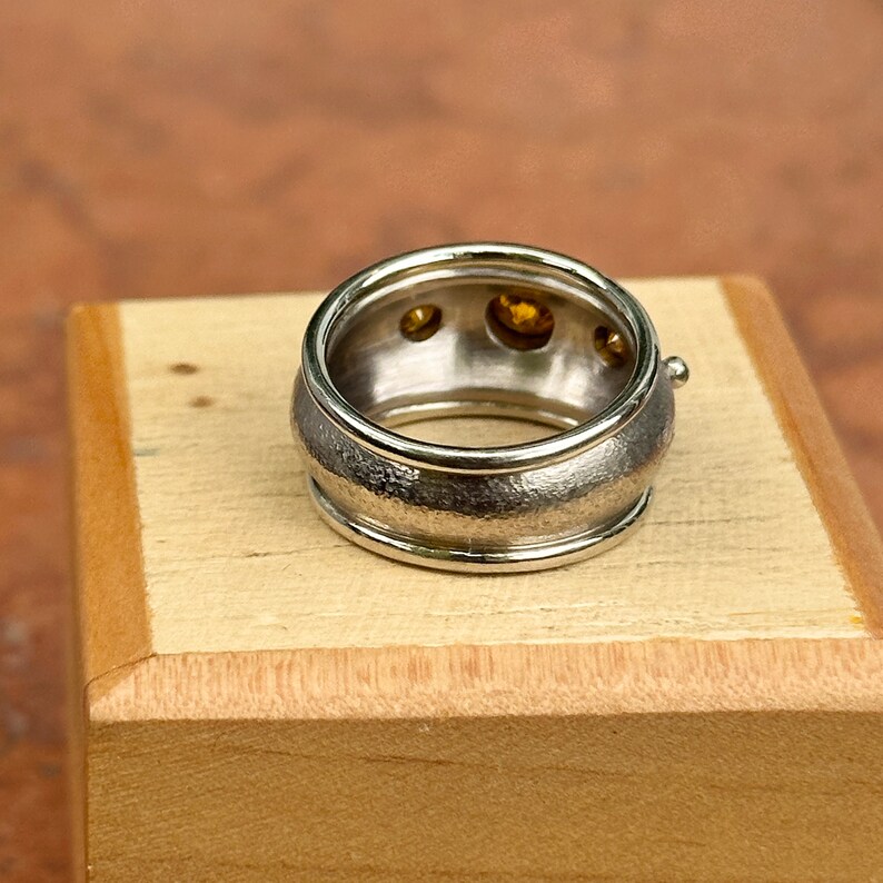 May include: A wide silver ring with three round, yellow gemstones set inside. The ring has a textured band and a smooth, polished interior. The ring is displayed on a small wooden block.