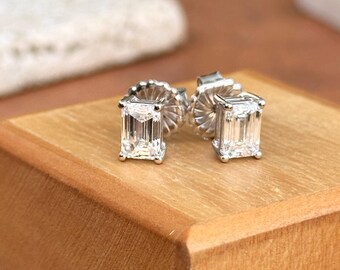 May include: A pair of square-cut, clear gemstone stud earrings. The earrings are set in a silver metal with a post and butterfly backing. The earrings are displayed on a wooden surface.