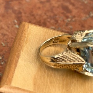 May include: Close-up of a gold ring with a large, light blue gemstone. The ring has an ornate, textured band and a detailed setting around the gemstone. The ring is set on a wooden surface.